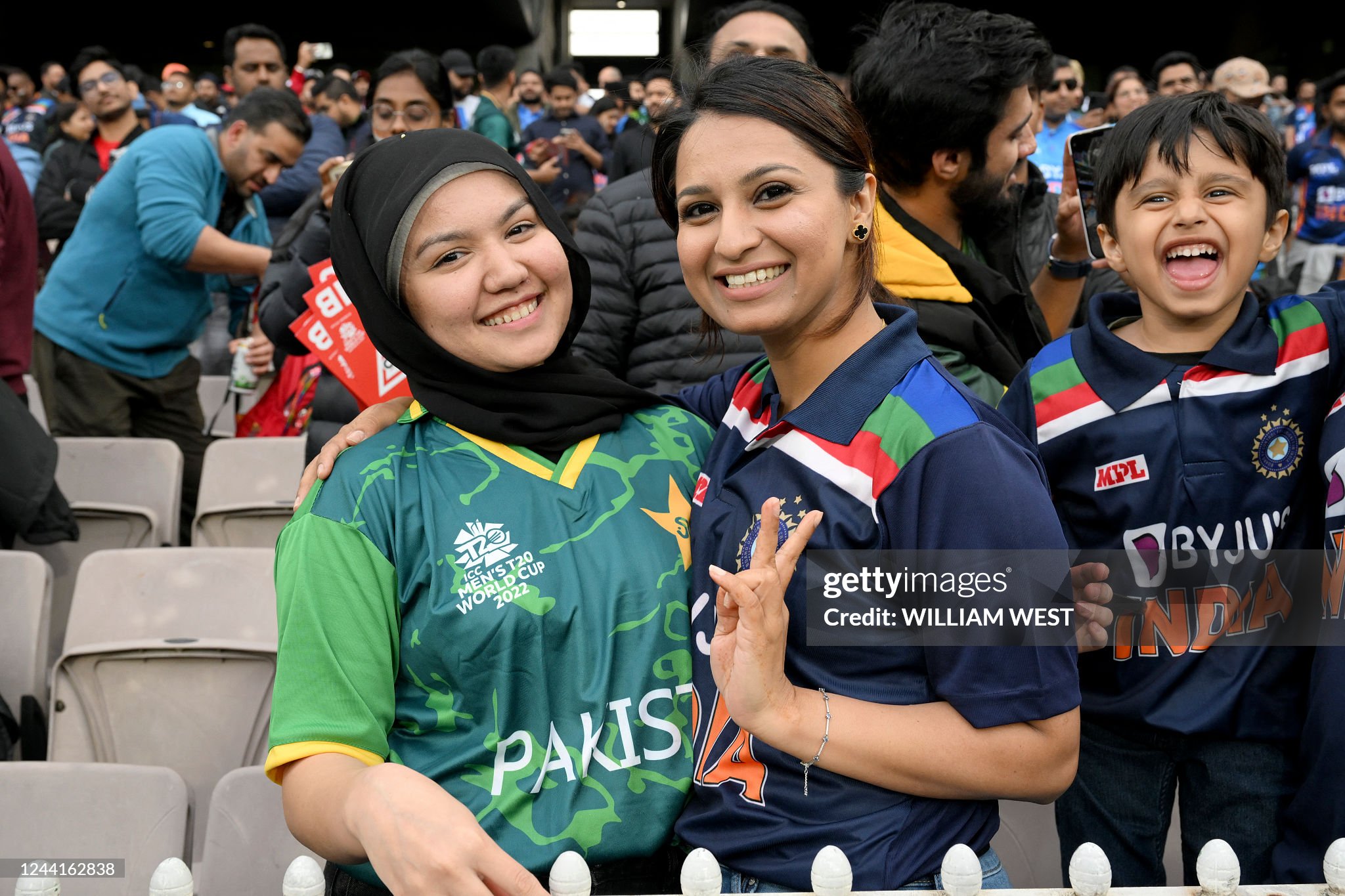 An Indian fan and a Pakistan fan smiling and posing together in team jerseys at the Melbourne Cricket Ground
